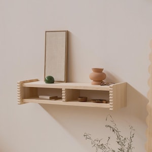 Op de afbeelding: Een lichtgekleurde houten plank met twee niveaus, met een ingelijst canvas, een groene bol, een kleine terracotta vaas en andere decoratieve items. De plank heeft een getextureerd randontwerp en is aan een neutraal gekleurde muur bevestigd.