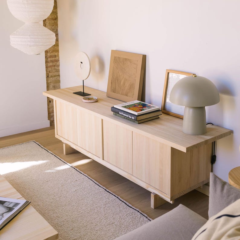 May include: A light wood cabinet with four doors and a light wood top. There are three books and a lamp on the top of the cabinet. The lamp is a mushroom-shaped lamp with a gray shade.
