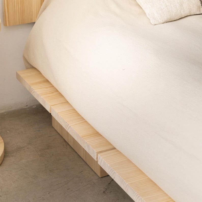 Puede incluir: Un marco de cama de madera de color claro con una base de plataforma. La cama est&aacute; parcialmente cubierta con una s&aacute;bana de color crema y una almohada. El marco es de madera clara y el dise&ntilde;o es minimalista.