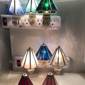 Picture of six lamp shade night lights plugged into outlets. They are blue, light blue, green, white, dark blue, and clear. There are two lamp shades on the table, one is red and one is pink.