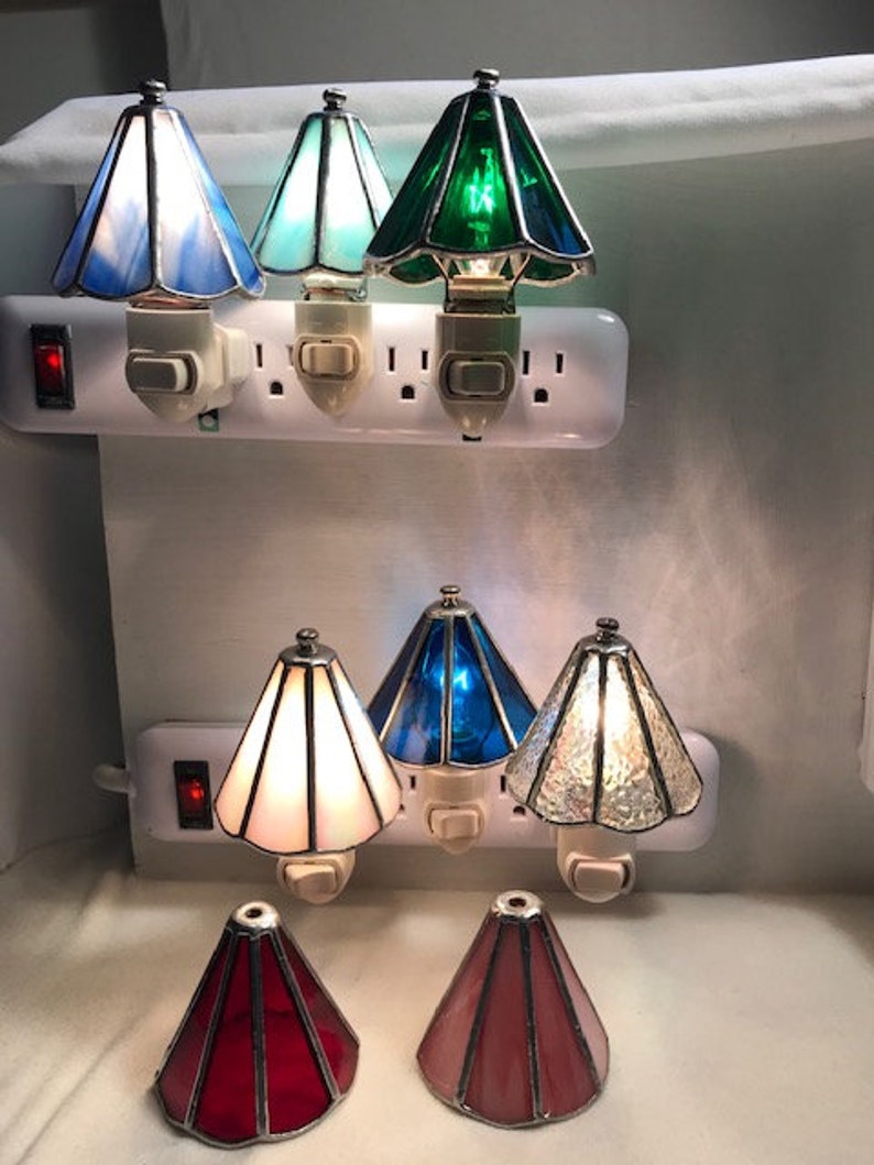 Picture of six lamp shade night lights plugged into outlets. They are blue, light blue, green, white, dark blue, and clear. There are two lamp shades on the table, one is red and one is pink.