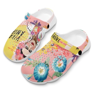 May include: A pair of white and pink floral print clogs with a yellow strap that says "Viva La Amore". The clogs have a white base and a pink and yellow floral print on the top. The strap is yellow and has the words "Viva La Amore" written on it in black.