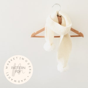May include: A white knitted scarf with pom poms on a wooden hanger. The scarf is hanging on a white wall. The image includes a logo that says "Handmade Modern Market Pattern PDF".