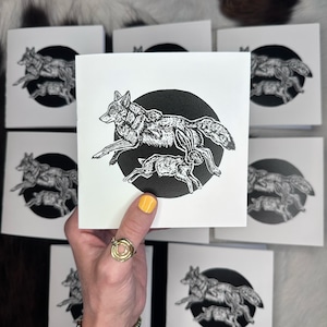 Puede incluir: Un conjunto de tarjetas cuadradas blancas con una ilustración en blanco y negro de un lobo y un animal más pequeño, posiblemente un conejo, corriendo dentro de un círculo negro. Las tarjetas son sostenidas por una mano con un anillo y una pulsera dorados.