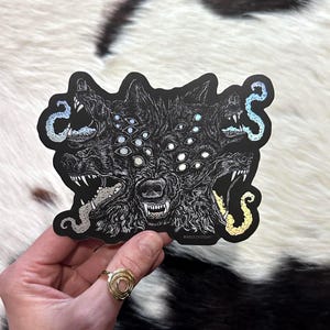 May include: A black sticker featuring a multi-headed wolf with multiple eyes and open mouths. The illustration is detailed with white and silver accents, and held by a hand. The sticker has a unique, artistic design.