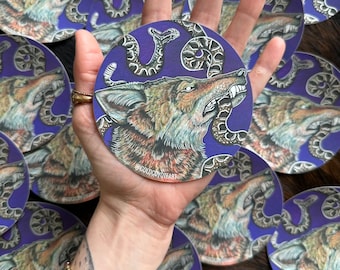 May include: A round sticker with a purple background features a detailed illustration of a wolf's head with a snake wrapped around it. The wolf is snarling with its mouth open, and the snake is coiled around its head and neck. The sticker has the text "@GOLDCQYDEART" in the bottom right corner.