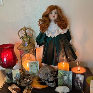 Puede incluir: Una muñeca con cabello rojo y un vestido verde se encuentra cerca de velas encendidas, cartas del tarot y una calavera decorativa. Un portavelas de vidrio rojo y una linterna dorada crean un tema místico.