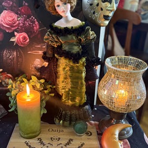 May include: A decorative scene featuring a vintage doll in a green dress, a lit candle, a Ouija board, and a golden mask. The Ouija board has the word "Ouija" and the alphabet. An orange crescent moon and a mosaic glass candle holder are also present.