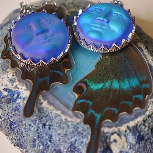 Könnte beinhalten: Ein Paar regenbogenfarbene blaue Glasmond-Ohrringe mit silbernen Einstellungen, die auf einem blauen und weißen Achat-Geoden mit einem Schmetterlingsflügel präsentiert werden.