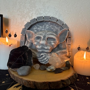 May include: A grey stone gargoyle sculpture with a stone archway behind it. The gargoyle is sitting on a wooden slice with various crystals and stones around it. There are two white candles on either side of the sculpture.