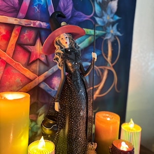 May include: A decorative figurine of a witch wearing a black dress and a red-brimmed hat, holding a broom and a cauldron. The figurine is surrounded by lit candles of various sizes and colors, with a colorful backdrop featuring stars and floral designs.