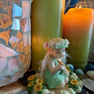 Pode incluir: Uma pequena estatueta de querubim com um vestido e chapéu verdes, segurando uma flor amarela. O querubim está rodeado por flores amarelas e está sentado sobre uma base em forma de nuvem. Velas altas verdes e laranja estão no fundo, junto com uma lâmpada de vidro de mosaico.