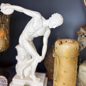 Könnte beinhalten: Eine weiße Marmorstatue eines Diskuswerfers in Bewegung. Die Skulptur steht auf einem weißen Sockel. Eine hohe, zylindrische Kerze mit goldenen Akzenten und einer Kordelbindung befindet sich im Hintergrund. Weitere dekorative Gegenstände sind ebenfalls zu sehen.