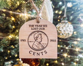 May include: Wooden Christmas tree ornament with the text "THE YEAR WE LOST OUR" and a penny design. The ornament includes the years 1793 and 2025, and the word "CENTS". The ornament is hanging on a Christmas tree.