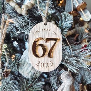 May include: Wooden Christmas tree ornament with the text "THE YEAR OF 67 2025". The numbers "67" are in gold, and the ornament is hanging on a Christmas tree.