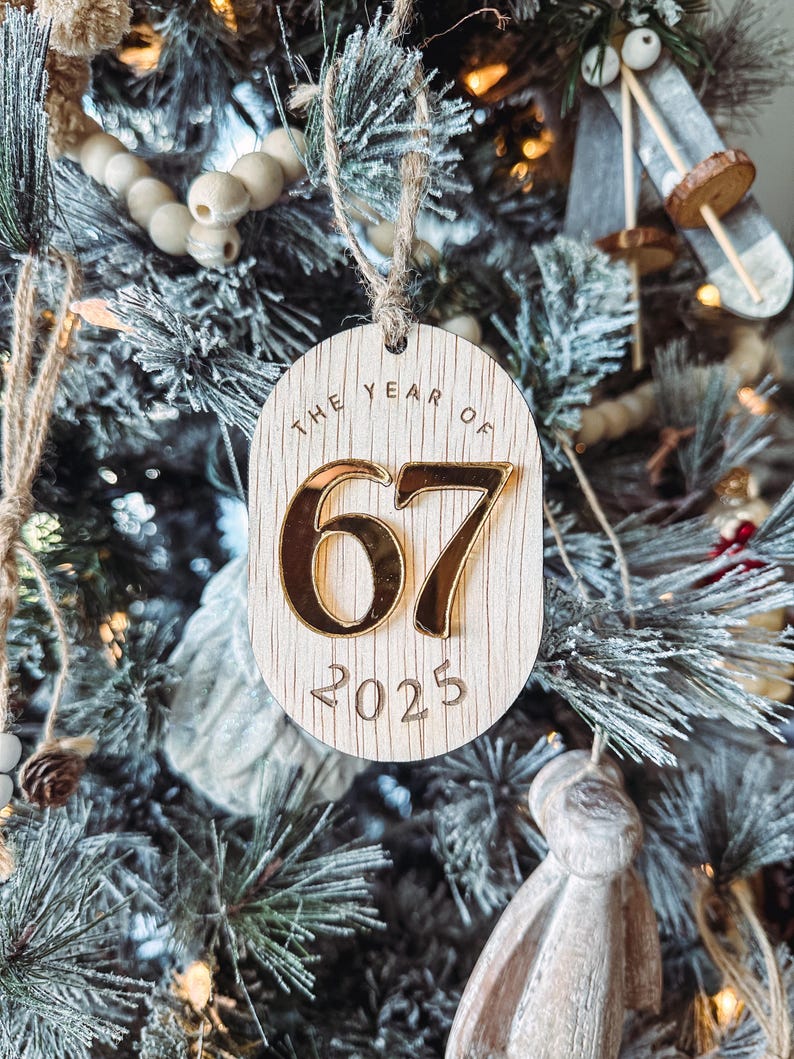 May include: Wooden Christmas tree ornament with the text "THE YEAR OF 67 2025". The numbers "67" are in gold, and the ornament is hanging on a Christmas tree.