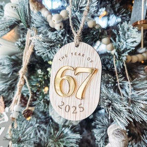 May include: Wooden Christmas ornament with the text "THE YEAR OF 67 2025". The ornament is oval-shaped with a light wood grain finish. The numbers "67" are gold, and the year "2025" is engraved below. The ornament hangs from a twine loop.