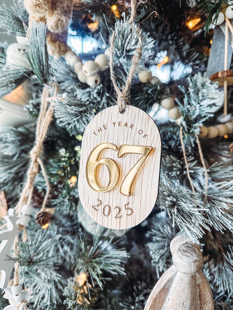 May include: Wooden Christmas ornament with the text "THE YEAR OF 67 2025". The ornament is oval-shaped with a light wood grain finish. The numbers "67" are gold, and the year "2025" is engraved below. The ornament hangs from a twine loop.