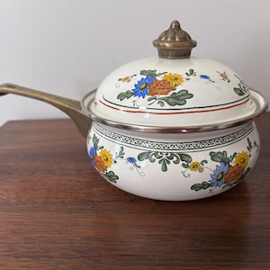 May include: A cream-colored enamel saucepan with a matching lid, featuring a floral pattern of blue, orange, and yellow flowers with green leaves. The pot has a brass-toned metal handle and a matching brass-toned knob on the lid.