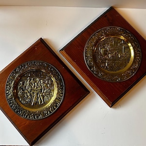 May include: Two square wooden wall plaques, each featuring a circular brass plate with embossed relief scenes. One plate depicts figures gathered around a table, while the other shows a horse-drawn carriage. Both have ornate, decorative borders.