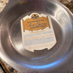 May include: A silver-toned metal pan with a central paper label featuring the text Revere Ware, Solid Copper/Stainless Steel, Paul Revere Signature Collection, and instructions for removing the protective coating before use.