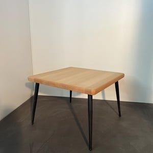 Op de afbeelding: Een vierkante tafel met een lichtgekleurd natuurlijk houten blad met afgeronde hoeken en vier slanke, matzwarte metalen poten die naar de vloer toe taps toelopen. Het tafeloppervlak toont een duidelijke houtnerf en een gladde rand.