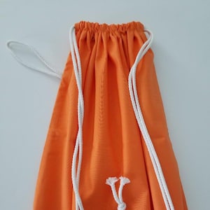 May include: An orange drawstring bag with white rope straps. The bag is made of a solid orange fabric and has a gathered top. The white rope forms the drawstring closure and shoulder straps.