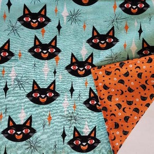 May include: A teal fabric with a repeating pattern of black cat faces with red eyes, orange mouths, and starburst and diamond shapes. A corner of orange fabric with a moon and star pattern is visible.