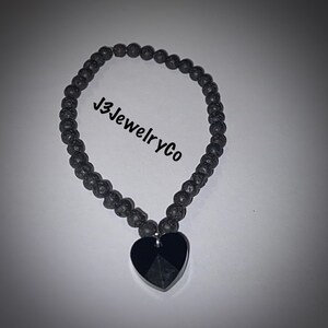 Può includere: Braccialetto con perline di roccia vulcanica nera con un ciondolo a forma di cuore in cristallo nero. Il braccialetto è infilato su un cordoncino nero e ha il logo J3JewelryCo.