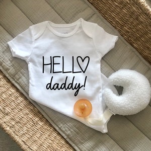 May include: White baby onesie with black text that reads "Hello daddy!" with a heart symbol. The onesie is on a woven surface with a white pacifier and a white stuffed toy.