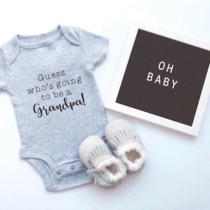 Peut inclure: Une grenouillère grise avec le texte "Devinez qui va être un grand-père !" Un tableau d'affichage blanc avec le texte "OH BABY" et une paire de chaussons de bébé blancs.