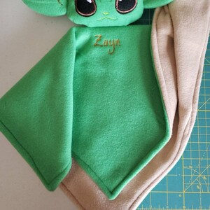 May include: Green and tan baby blanket with a plush embroidered character of a green alien with large eyes. The blanket has the name "Zayn" embroidered on it.