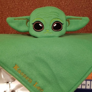 May include: Green plush toy of a character with large eyes and ears, sitting on a green fleece blanket with the name "Kassen Lee" embroidered in yellow thread.