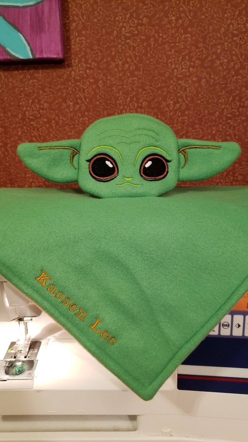 May include: Green plush toy of a character with large eyes and ears, sitting on a green fleece blanket with the name "Kassen Lee" embroidered in yellow thread.