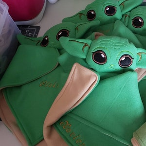May include: Four green plush toys with large eyes and pointy ears, resembling a popular fictional character. Each toy is lying on a tan and green blanket with a name embroidered on it. The names are Eli, Xavier, and two others that are not visible.