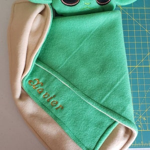 May include: A green and beige baby blanket with a plush green character with large eyes and ears. The blanket has the name "Javier" embroidered on it in gold thread.