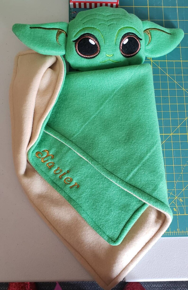 May include: A green and beige baby blanket with a plush green character with large eyes and ears. The blanket has the name "Javier" embroidered on it in gold thread.
