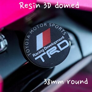 Puede incluir: Una pegatina redonda de resina 3D abovedada de 38 mm con un patrón de fibra de carbono en blanco y negro. La pegatina presenta el logotipo de Toyota Motorsports, el texto "Racing Development" y el logotipo de TRD en blanco.