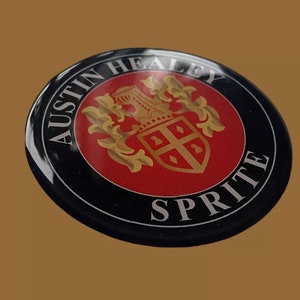 May include: A black and red round emblem with the text "Austin Healey Sprite" in white lettering. The emblem features a gold crest with a red shield and a crown.