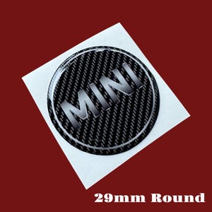 May include: A round, black and silver MINI emblem with a carbon fiber pattern background. The word "MINI" is in silver, and the sticker is 29mm in diameter. The emblem is on a white square, set against a dark red background.