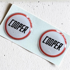 Puede incluir: Dos pegatinas redondas blancas con un círculo rojo alrededor del borde. La palabra "COOPER" está impresa en letras negras dentro del círculo.