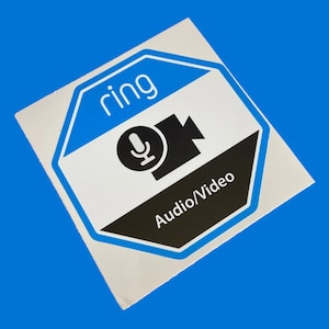 Puede incluir: Una pegatina octogonal blanca y azul con la palabra "ring" en texto azul. La pegatina tiene un icono en blanco y negro de un micrófono y una cámara de video con el texto "Audio/Video" en texto blanco.