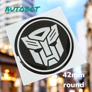 May include: A black and white round sticker with the Autobot logo from the Transformers franchise. The sticker is 42 mm in diameter and says "Autobot" and "42mm round".