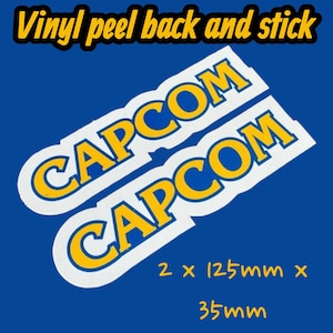 May include: Two white vinyl peel-and-stick decals with the word "CAPCOM" in yellow and blue. The decals are 12.5cm x 3.5cm each. The text "Vinyl peel back and stick" is at the top of the image, in yellow.