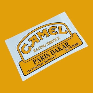 Può includere: Un adesivo rettangolare con sfondo bianco e logo giallo e marrone. Il logo presenta la parola "CAMEL" in un font stilizzato, con le parole "RACING SERVICE" sottostanti. Sotto il logo ci sono le parole "PARIS DAKAR" e "CITROËN SPORT".