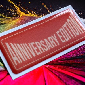Op de afbeelding: Een rode, rechthoekige sticker met een witte rand en de tekst "Anniversary Edition" in witte letters.