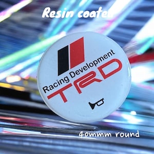 Könnte beinhalten: Ein weißer, runder, harzbeschichteter Knopf mit dem Text "Racing Development TRD" in rot und schwarzer Schrift. Der Knopf hat außerdem ein kleines, schwarzes, stilisiertes Bild eines Horns. Der Knopf hat einen Durchmesser von 40 mm.