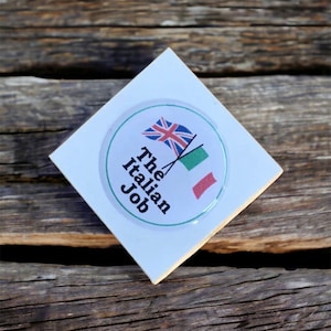 May include: A white square sticker with a round, clear, raised sticker on top. The round sticker has a green and red Italian flag and a Union Jack flag crossed over each other. The text "The Italian Job" is written in black on the round sticker.