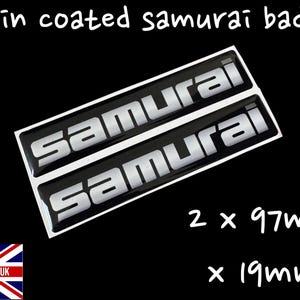 Pode incluir: Duas insígnias "Samurai" pretas revestidas a resina com letras prateadas. Cada insígnia mede 9,7 cm x 1,9 cm. O texto "Resin coated samurai badges" está no topo. A bandeira Union Jack com "MADE IN THE UK" está na parte inferior.