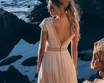May include: A woman wearing a white wedding dress with a lace bodice and a flowing skirt stands on a rocky cliff overlooking the sea.